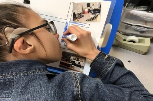 A young girl wearing a cochlear implant and glasses drawing