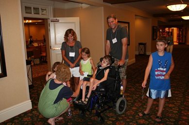 image of 4 children 3 girls and one boy.  One girl is in a wheel chair with a boy and girl standing next to her. A woman stands in front of a door and a man is behind the wheelchair.  Another woman is crouching facing the others and hugging the other girl.