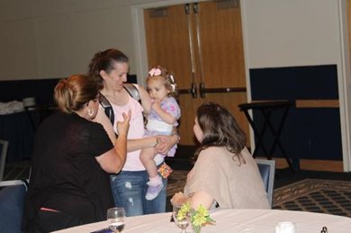 image of a woman holding a baby while a woman is standing in front of the baby while another woman is seated at the table looking at the interaction.