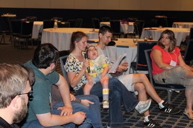 image of a group of seated people who appear to be listening to a woman reading off a piece of paper with a child on her lap